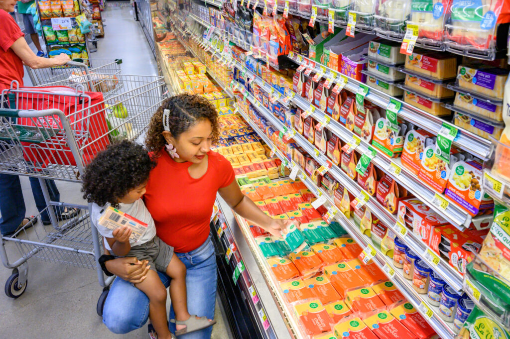 Una mujer con un niño en brazos selecciona queso en un estante de un supermercado, rodeada de una variedad de quesos envasados. Se ve a otro comprador al fondo.