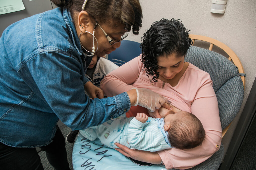 A mother breastfeeds her baby while a woman in a denim shirt assists, both seated in a room.