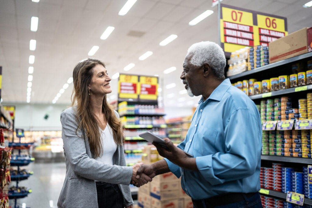 Two people are shaking hands in a supermarket aisle. Shelves are stocked with various products, and signs with numbers are visible above.