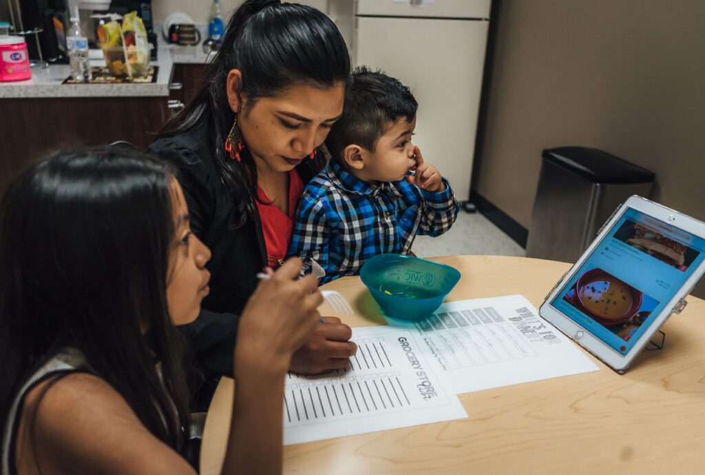 A woman and two children sit at a table with a tablet displaying informative recipes, grocery lists, and a bowl of snacks.