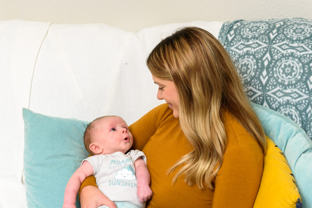 A woman with long hair gazes lovingly at a baby in her arms, seated on a couch adorned with blue and yellow cushions and a patterned blanket, reminiscent of the warmth found in Louisiana WIC-supported homes.