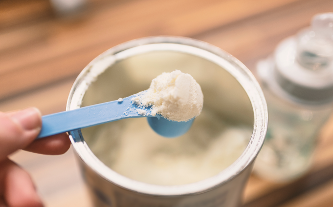 A hand holds a blue scoop with white powdered formula, perfect for infants aged 6-12 months, over an open container. A bottle is visible in the background.