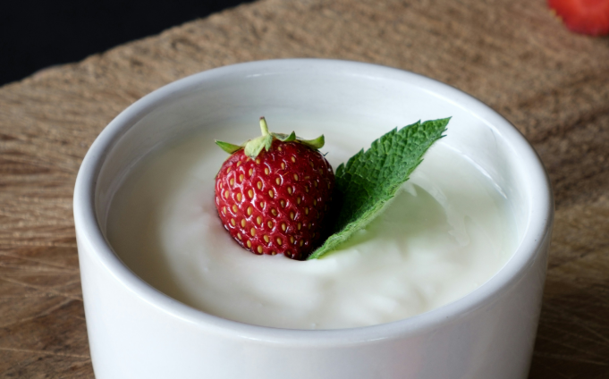 A white bowl filled with plain yogurt, topped with a whole strawberry and a fresh mint leaf, placed on a wooden surface.