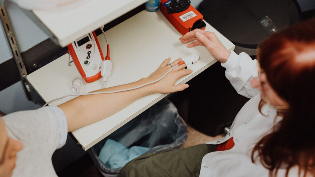 A medical provider in a lab coat gently places a pulse oximeter on the fingertip of another person, who sits beside a table brimming with medical resources.