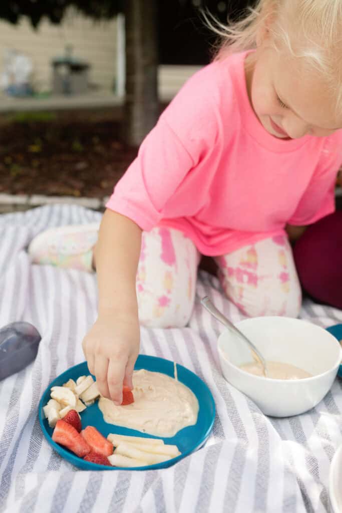 A young child enjoys a strawberry slice with creamy dip, promoting child nutrition, while sitting on a striped blanket outdoors.