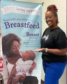 A woman stands next to a banner promoting breastfeeding and peer support, featuring a mother with her baby and info about breast pumps.
