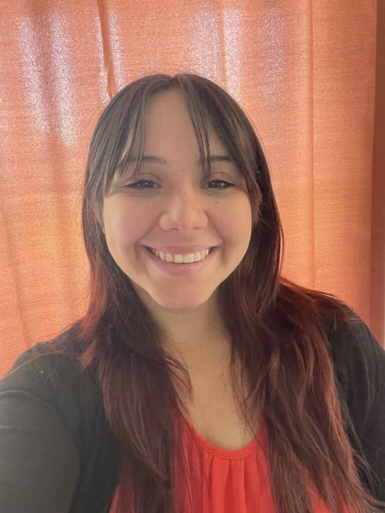 A Breastfeeding Peer Counselor with long brown hair smiles at the camera, standing before orange curtains in October 2025.
